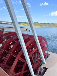 The image shows the side view of a red paddle wheel of a boat, with a backdrop of a blue sky and a distant shoreline.