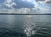 The image shows a serene lake with glistening water reflecting the sunlight, a treelined horizon, and a partly cloudy sky above.