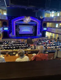 The image shows an audience seated at tables inside a venue, looking towards a stage with a decorative arch that reads