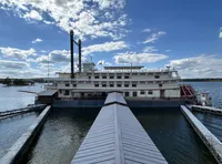 The image displays a large, multi-deck riverboat with a traditional paddlewheel, docked at a pier with a walkway leading up to it under a partly cloudy sky.