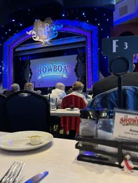 The image shows an audience seated at dining tables inside a theater, waiting for a performance to begin at the