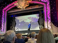 A man in a blue suit stands onstage before an audience with a backdrop of a cityscape on the screen behind him.