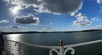 The image captures a panoramic view of a serene lake with a glittering sun above, clouds scattered across the sky, and a partial view of a person beside a white fence.