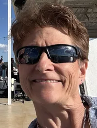 Two people are smiling for a selfie on a sunny day with a clear sky, some clouds in the background, and an outdoor venue with string lights overhead.