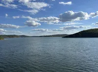 The image shows a tranquil expansive lake with hills in the background under a partly cloudy sky.