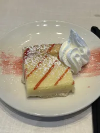 A slice of cheesecake with a dollop of whipped cream is served on a white plate, garnished with powdered sugar and a drizzle of strawberry sauce.