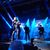 A live music performance shows several singers on stage with vibrant blue stage lighting and a captivated audience in the foreground.