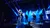 A live music performance shows several singers on stage with vibrant blue stage lighting and a captivated audience in the foreground.