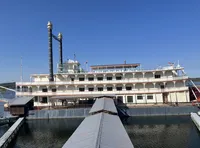 The image shows a large, multi-decked riverboat with traditional paddle-wheel design, moored at a dock with a long gangway on a clear day.