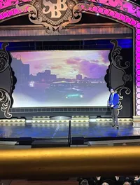 A person stands on stage with a large screen in the background displaying a cityscape at dusk, within a lavishly decorated theater setting.