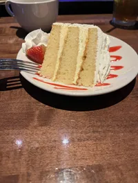 A slice of layered vanilla cake with white frosting, garnished with a strawberry and decorative sauce, is served on a white plate with a fork on the side, accompanied by a cup and glass on a wooden table.