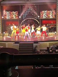 Performers in colorful costumes are dancing on stage during a live show with a band playing in the background, in front of an audience seated at tables.