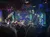 A live band performs on stage with colorful lighting and a starry backdrop as an audience watches from the foreground.