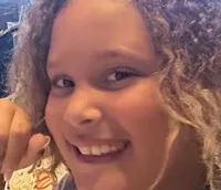 A smiling young person with curly hair sits at a table with a slice of cake, surrounded by people in a dimly lit function room.