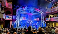 A lively musical performance is underway on a patriotically-themed stage with an audience in the foreground enjoying the show.