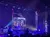 An illuminated stage scene with purple stage lights and a musician playing drums inside a transparent cage-like structure.