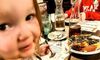 A child is smiling at the camera while sitting at a restaurant table with various dishes, including a dessert, in front of them.