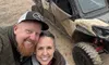 A smiling couple takes a selfie in front of muddy off-road vehicles, with a construction vehicle visible in the misty background.