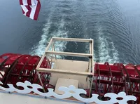 The image shows the stern of a paddlewheel steamboat with red paddlewheels churning water as it moves forward, accompanied by a waving flag.