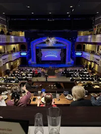 The image shows an audience seated at tables in a theater setting, enjoying a meal while watching a show called