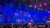 The image shows a vibrant theater set with an ornate, colorful backdrop depicting an exotic cityscape under a blue light, while audience members settle into their seats.