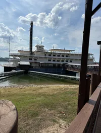 A large paddlewheel riverboat is docked by a grassy shore under a partly cloudy sky.
