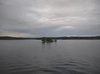 A small, tree-covered island sits in the middle of a calm lake under a cloudy sky with distant hills on the horizon.