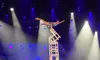A performer balances on a tall stack of chairs in a spotlight on stage.