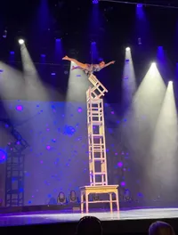 A performer balances on a tall stack of chairs in a spotlight on stage.