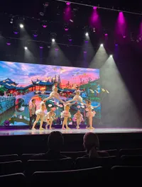 Performers are on stage in colorful costumes, balancing plates while an audience watches against a vibrant backdrop of a scenic landscape.