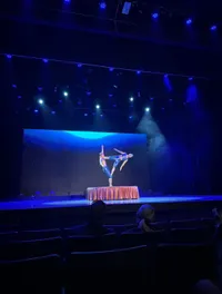 Two acrobats perform a balancing act on a small platform on a stage under dramatic lighting.