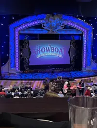 The image depicts an audience gathered in a theater for a performance titled