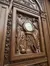 This image shows an ornate wooden carving featuring two figures flanking a central clock, surrounded by intricate decorative elements.