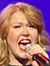 A woman is energetically singing on stage, holding a microphone, with vibrant purple lights in the background.