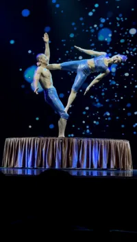 Two acrobats in blue outfits perform a balancing act on a circular platform against a backdrop of floating blue lights.