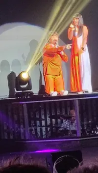 A performer in an orange outfit is kneeling on stage next to another person in a red and white dress, under bright yellow stage lights, with a drummer visible behind them.