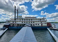 A large riverboat with two prominent smokestacks is docked at a pier under a partly cloudy sky.