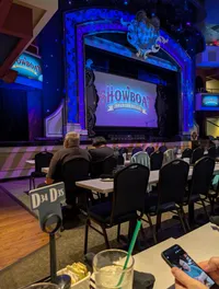 Audience members are seated at tables in a theater, facing the stage displaying a