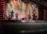 A lively band performs on stage under colorful lighting, with a singer, violinist, guitarist, and drummer.