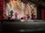 A lively band performs on stage under colorful lighting, with a singer, violinist, guitarist, and drummer.