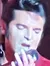 A performer in a red jacket is passionately singing on stage, surrounded by a band with a guitarist and a singer.