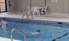 A person is swimming in an indoor pool with a white poolside chair and table nearby.