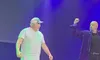Two men are on stage, one wearing a striped shirt and cap, while the other raises his fist, under blue and green lighting.