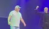 Two men are on stage, one wearing a striped shirt and cap, while the other raises his fist, under blue and green lighting.