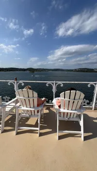 Two people are relaxing in Adirondack chairs on a deck overlooking a scenic lake with a partly cloudy sky.