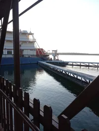 A riverboat with a prominent red paddle wheel is docked at a pier on a calm body of water beside a wooden fence.