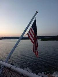 An American flag is flying on a pole over a body of water at dusk, with a scenic view of distant hills.
