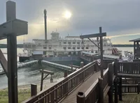 A large paddleboat docked at a pier is viewed from a wooden walkway as the sun sets in the background.