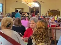 The image shows a crowded indoor dining area with people seated at tables, a chandelier hanging from the ceiling, and stage lighting equipment above.