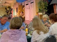 Several people are seated at tables with plates of food, facing a stage set designed like a rustic outdoor scene with an 